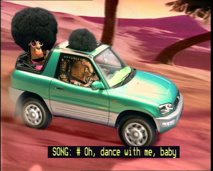 Example of captioned lyrics: frame from movie showing animated camels with afros singing in a car with captions: 'SONG: #Oh, dance with me, baby'