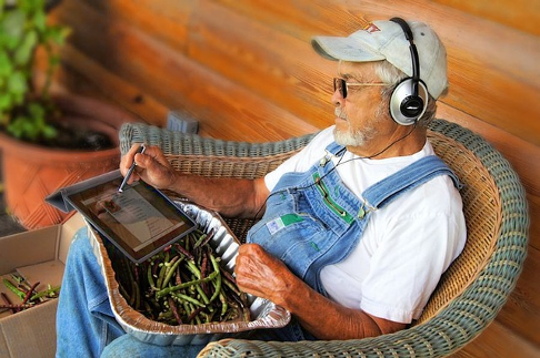 Elderly farmer wearing headphones whilst using a stylus on a tablet device, over an aluminum tray of peas resting on his lap
